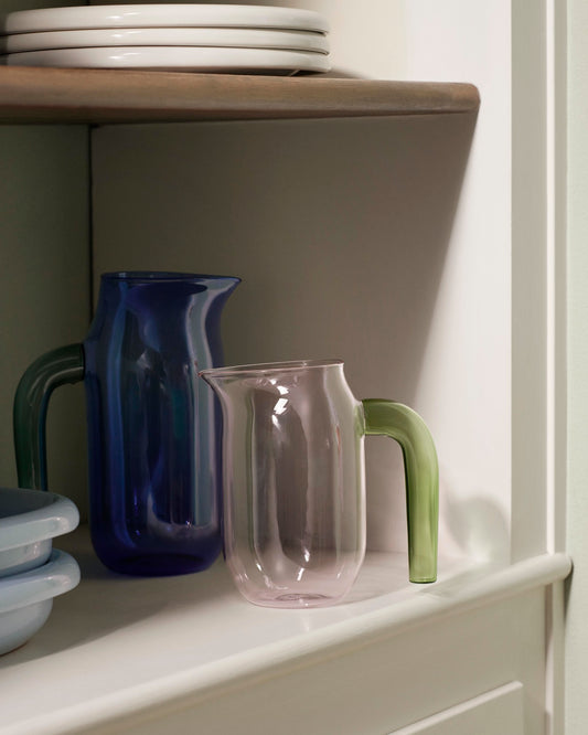 A lifestyle shot of the small pink HAY borosilicate glass jug designed by Jochen Holz, highlighting its distinctive asymmetrical rim and downward-curved handle.