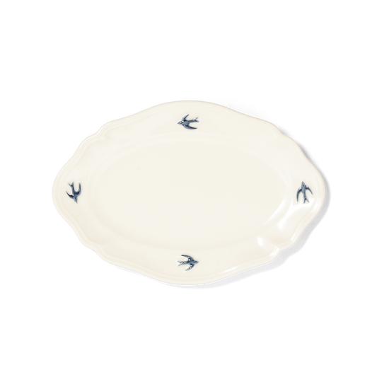 Studio M' Early Bird Oval Plate L in white porcelain, featuring a scalloped rim and a hand-painted blue swallow relief. A shot taken from the aerial angle.