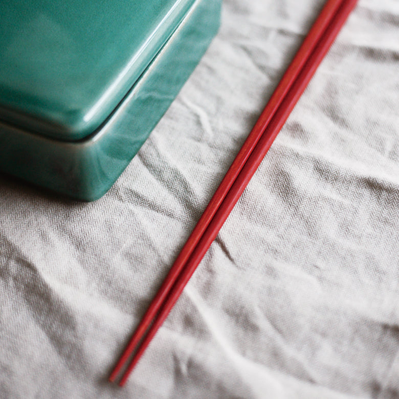 A lifestyle photo of Studio M' Lacquered Chopsticks featuring a sleek, minimalist profile and a subtle matte vermilion wipe-lacquer finish over natural wood grain.