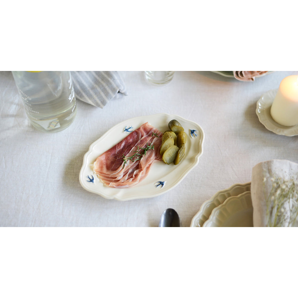 Studio M' Early Bird Oval Plate L in white porcelain, featuring a scalloped rim and a hand-painted blue swallow relief, on a table setting. 