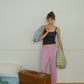 A woman stands in a minimalist bedroom setting, leaning against a white wall, holding two Hotel827 cotton travel pouches in different sizes and colours.