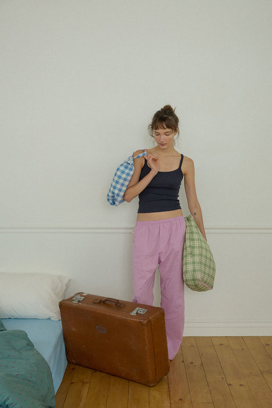 A woman stands in a minimalist bedroom setting, leaning against a white wall, holding two Hotel827 cotton travel pouches in different sizes and colours.