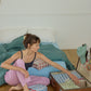 A woman in pink pajamas sits on a wooden floor next to an open vintage suitcase, packing Hotel827 cotton travel pouches.