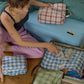 A woman packing Hotel827 cotton travel pocuhes in a bedroom setting, with several plaid toiletry pouches and a suitcase visible on the floor and bed.