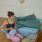 A woman kneels on a wooden floor beside a bed, packing different Hotel827 cotton travel pouches while an open brown suitcase lies nearby.