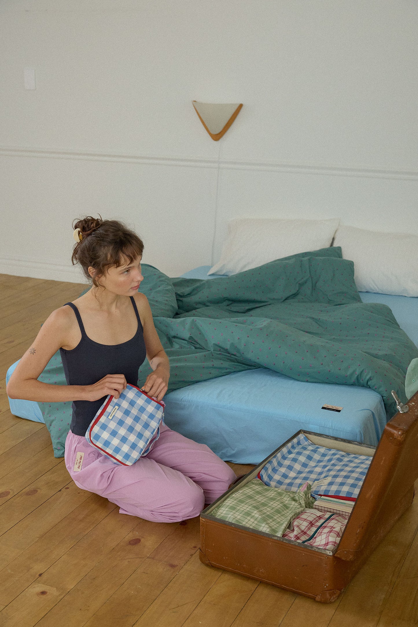 A woman kneels on a wooden floor beside a bed, packing different Hotel827 cotton travel pouches while an open brown suitcase lies nearby.