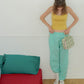 A woman stands in a bedroom wearing a yellow ribbed tank top and mint green baggy sweatpants, holding Hotel827 small cotton travel pouch in evergreen.