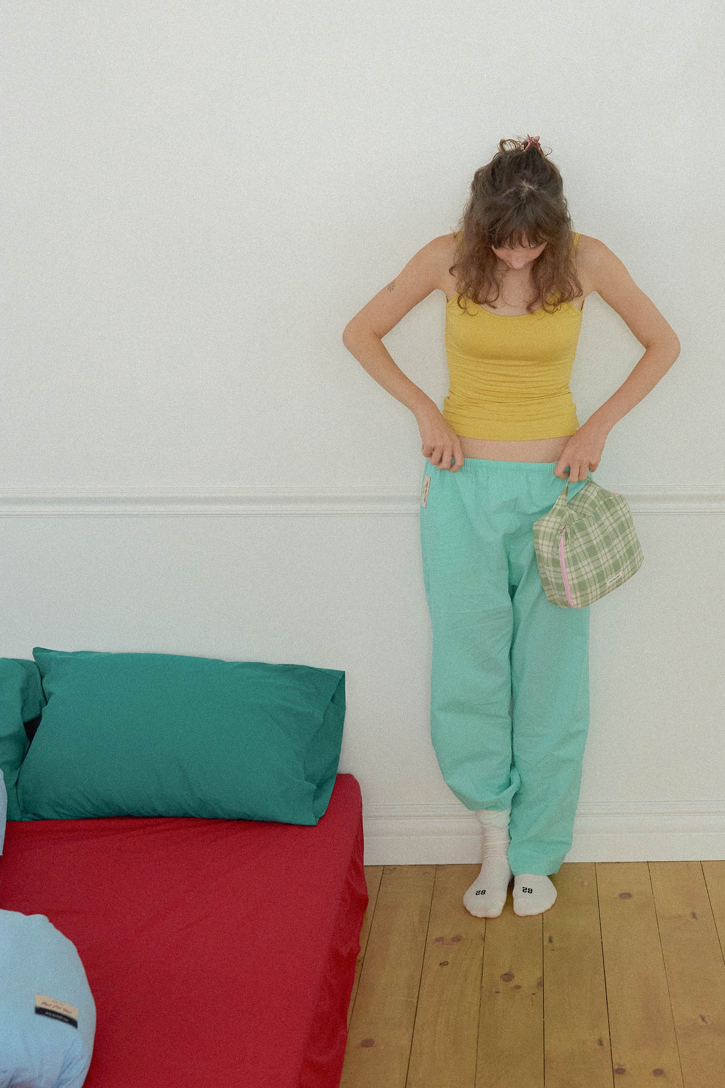 A woman stands in a bedroom wearing a yellow ribbed tank top and mint green baggy sweatpants, holding Hotel827 small cotton travel pouch in evergreen.