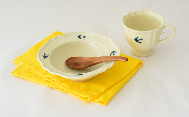 Studio M' Early Bird 170 porcelain bowl featuring a hand-painted blue swallow relief on a scalloped floral rim, paired with Early Bird Mug on a white dinning table. 