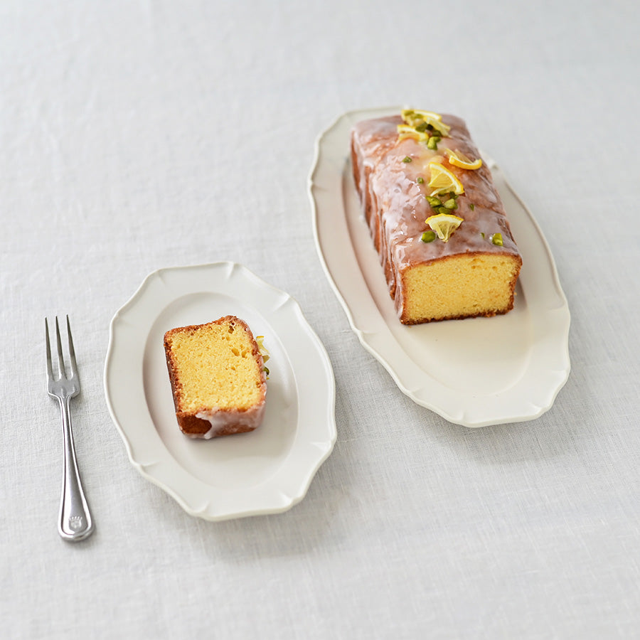 Studio M Ornata matte white 20cm oval plate with pastry and vintage dessert fork next to a Ornata Oval Plate 30cm on a white table cloth.
