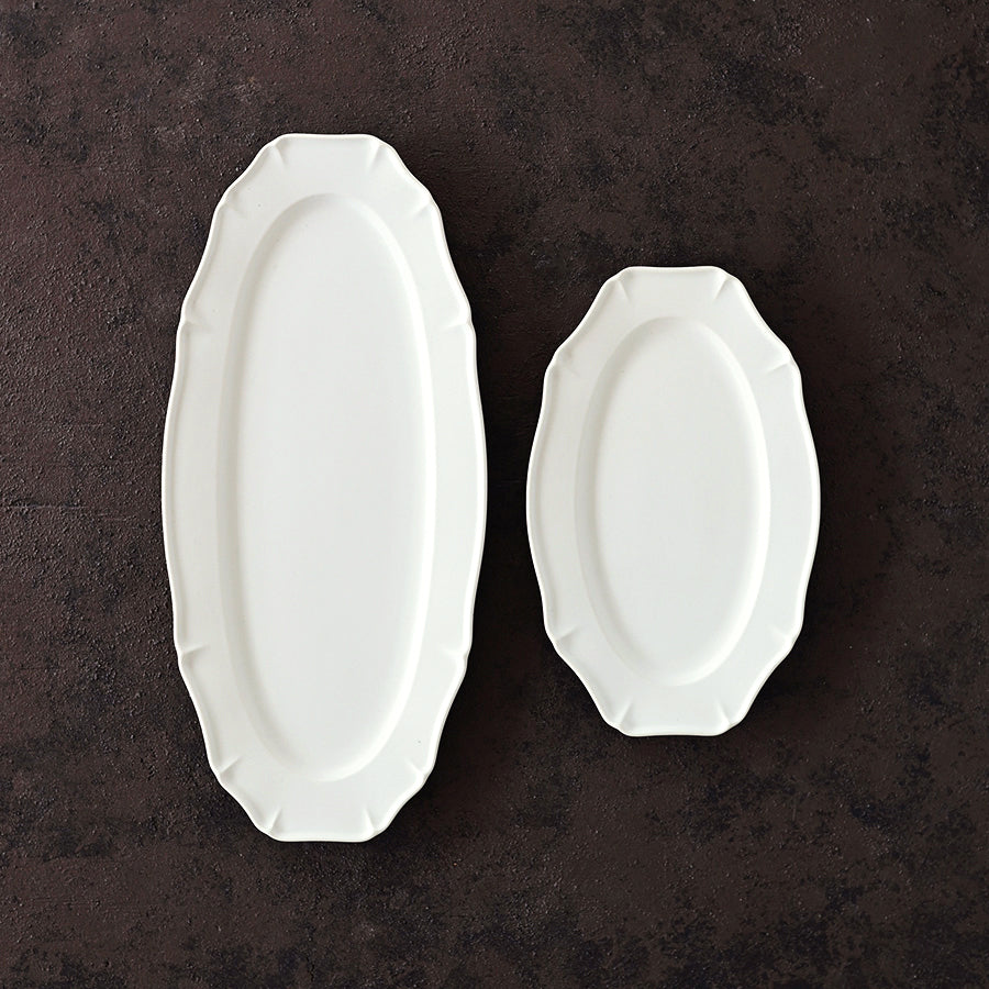 A comparison photo of Studio M Ornata matte white 20cm and 30cm oval plates on a brown metal surface. 