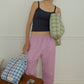 A woman stands indoors against a white wall wearing a dark navy camisole and loose light pink pants, holding two Hotel827 cotton travel pouches in different sizes and colours. 