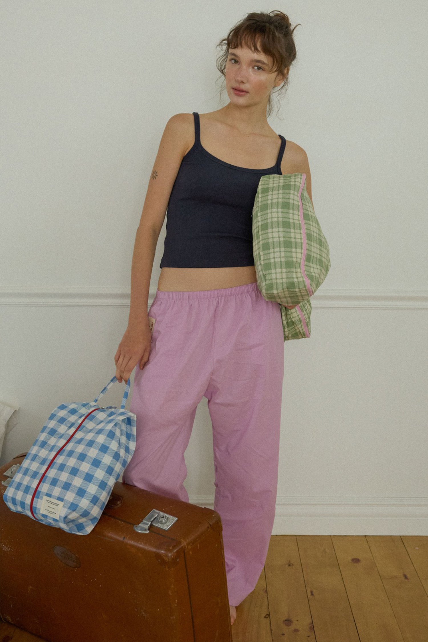 A woman stands indoors against a white wall wearing a dark navy camisole and loose light pink pants, holding two Hotel827 cotton travel pouches in different sizes and colours. 