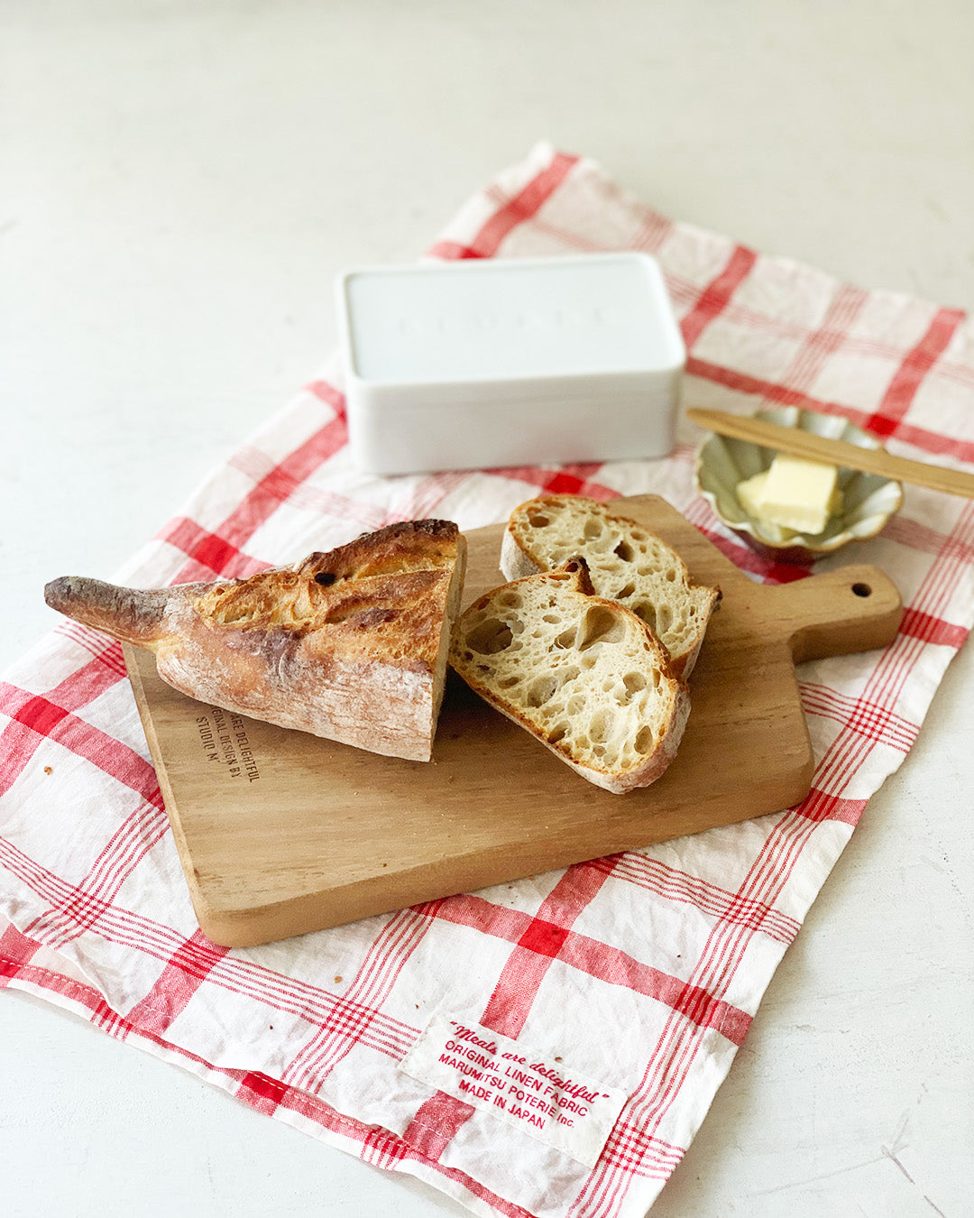 Studio M Japanese solid walnut cutting board collection in oblong medium size, serving cut baguette on a tea towel. 