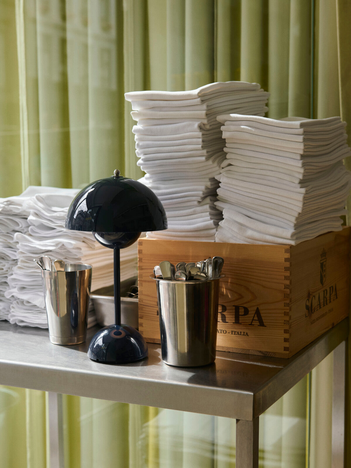 &Tradition Verner Panton Flowerpot VP9 Portable Table Lamp in Steel Blue on a stainless steel shelving next to napkins and cutlery.