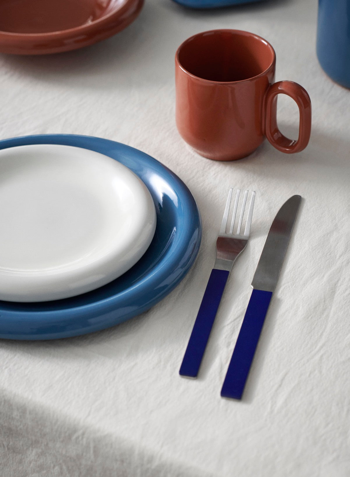 Dinnerware set with Hay Barro Plate 18cm in Off-White stacked on top of the Barro Plate 24cm in Dark Blue, red mug, fork, and knife on a white table cloth.