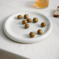 HAY Barro Plate 18cm in Off-White with green olives on a light surface with a glass of juice and bread.