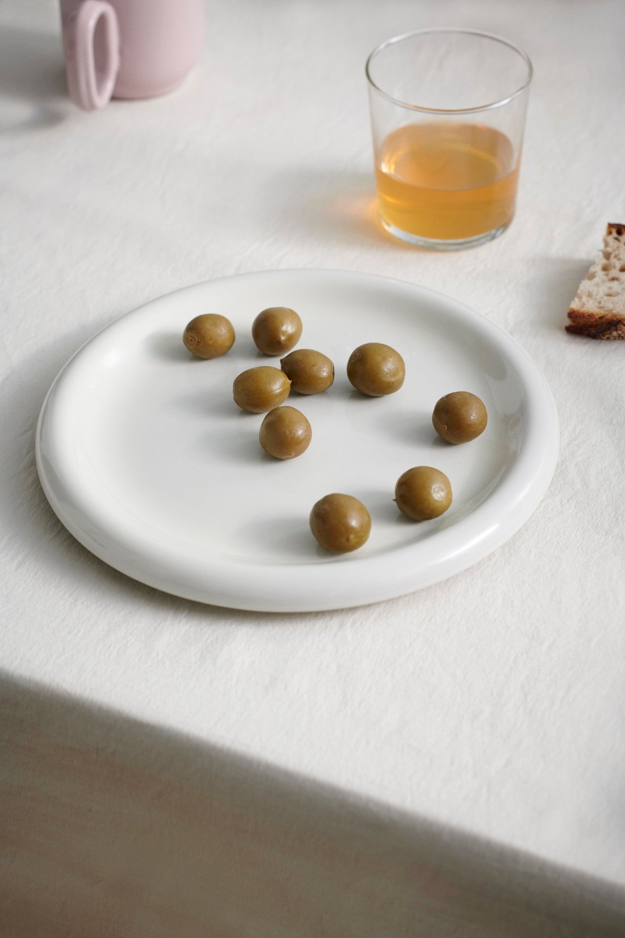 HAY Barro Plate 18cm in Off-White with green olives on a light surface with a glass of juice and bread.