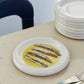 Hay Barro Plate 18cm in off-white with anchovies in a yellow sauce on a beige table with a stack of plates and cutlery.