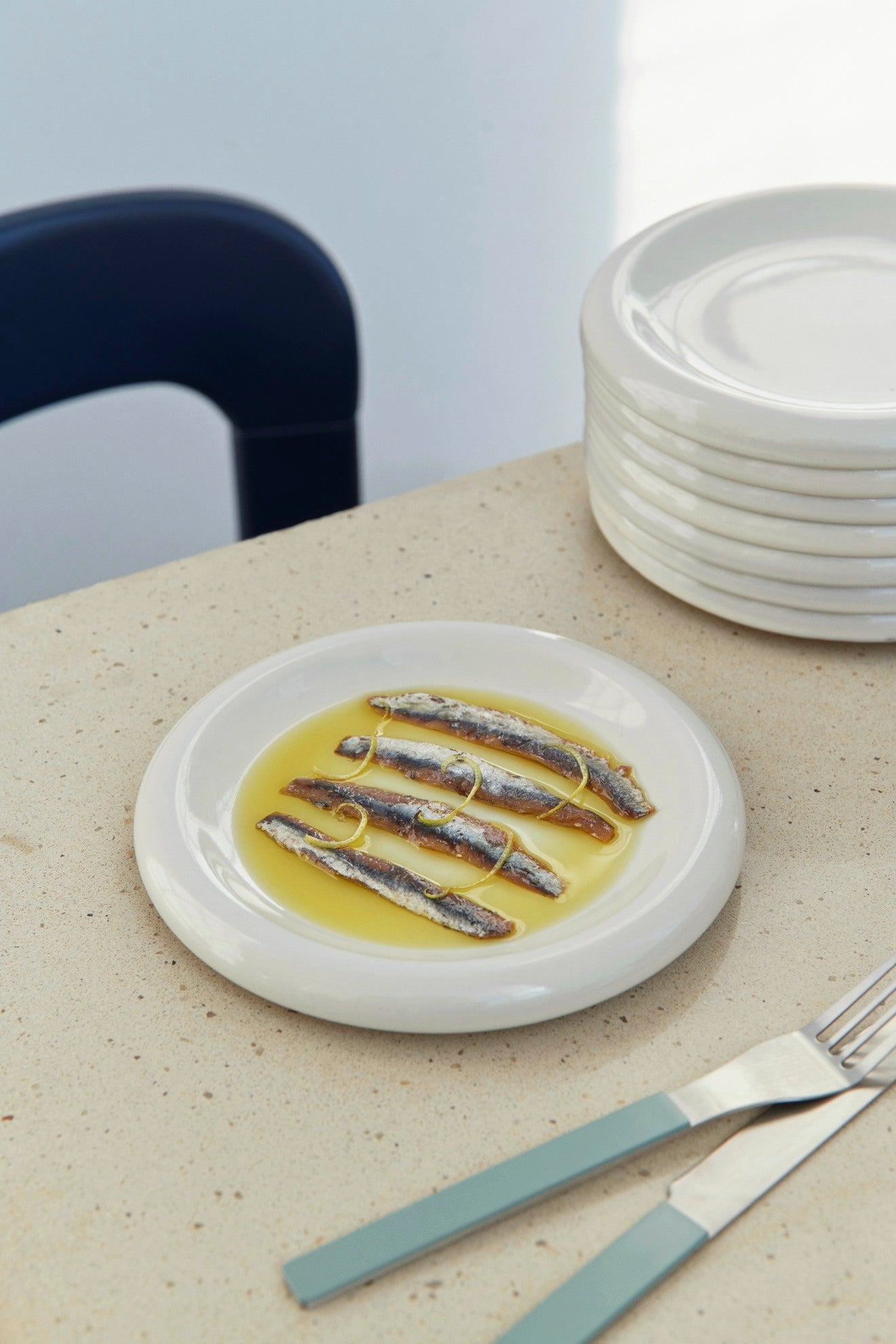 Hay Barro Plate 18cm in off-white with anchovies in a yellow sauce on a beige table with a stack of plates and cutlery.