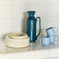 Stacked Hay Barro plates in off-white in twos sizes next to a few blue mugs and turquoise pitcher on a tiled kitchen counter