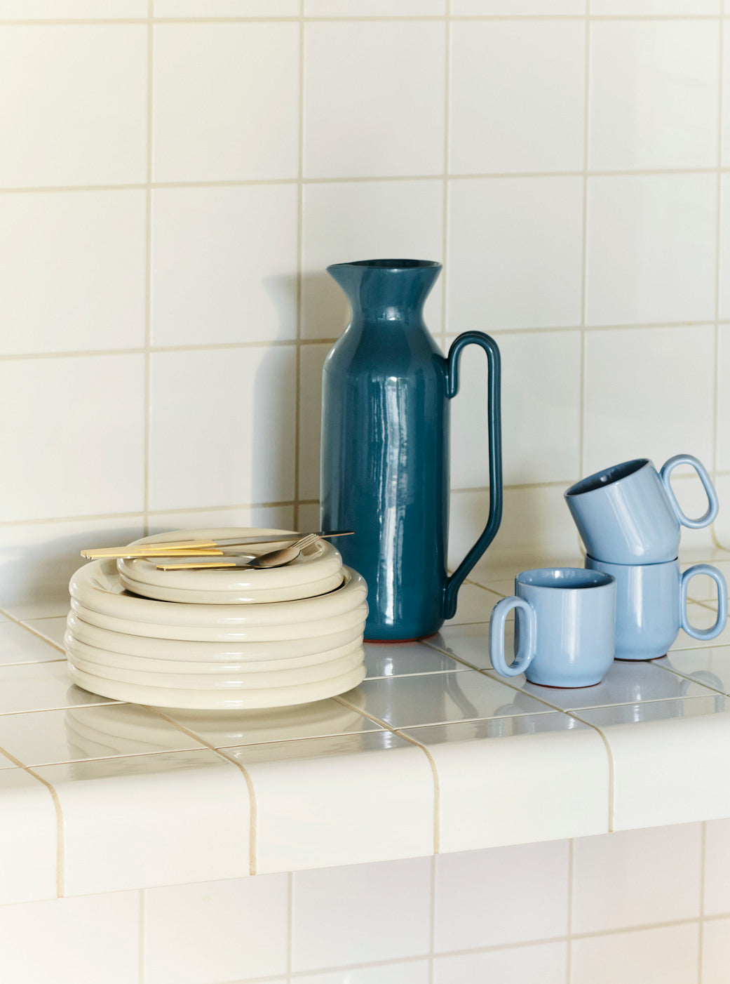 Stacked Hay Barro plates in off-white in twos sizes next to a few blue mugs and turquoise pitcher on a tiled kitchen counter