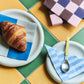 Two sizes of Hay Barro plates in offi-white with croissants and checkered napkins on a colorful tiled table.