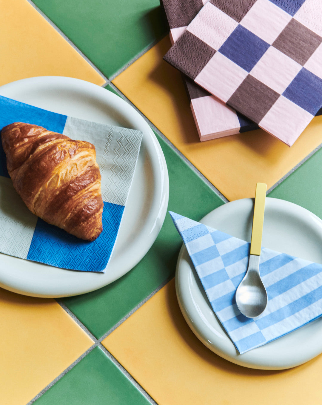Two sizes of Hay Barro plates in offi-white with croissants and checkered napkins on a colorful tiled table.