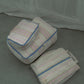 Three striped Hotel827 cotton travel pouches in Lilac, arranged on a green carpet in front of a green curtain backdrop.