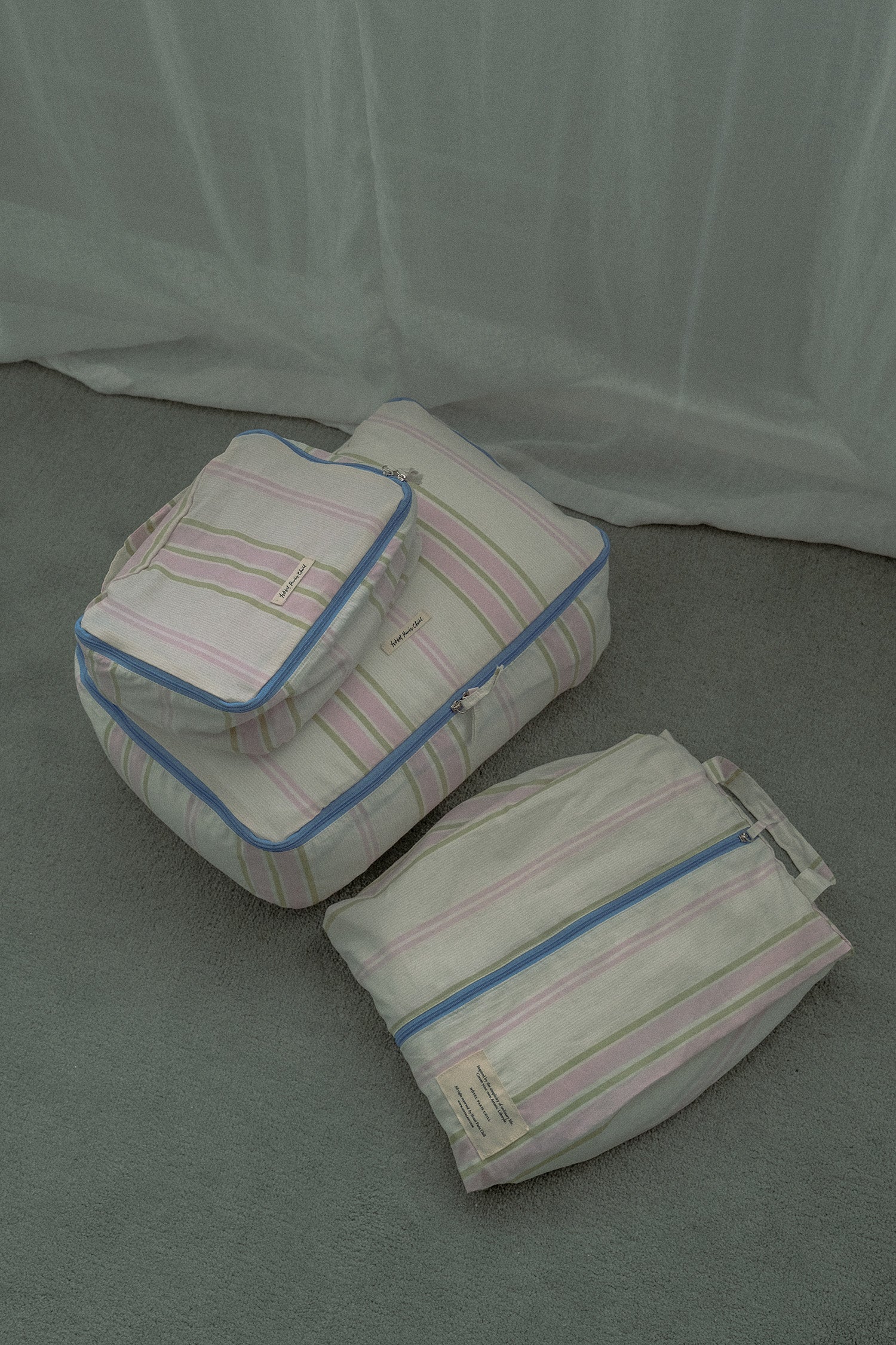 Three striped Hotel827 cotton travel pouches in Lilac, arranged on a green carpet in front of a green curtain backdrop.