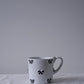Carron Paris white ceramic Noeud Noeud mug with a handle, decorated with small black bow motifs, resting on a textured white fabric surface.