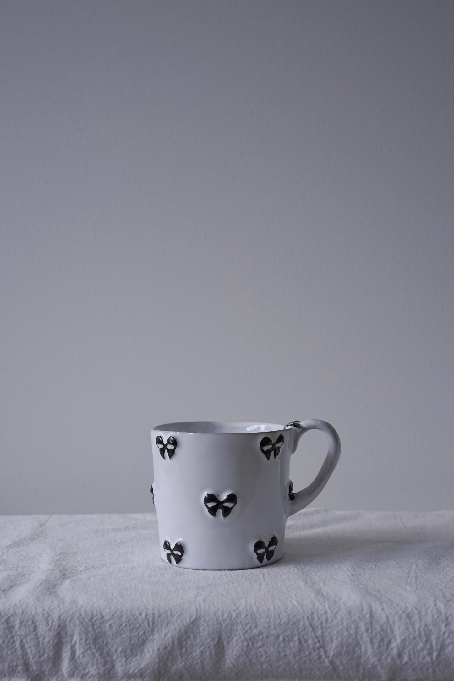 Carron Paris white ceramic Noeud Noeud mug with a handle, decorated with small black bow motifs, resting on a textured white fabric surface.