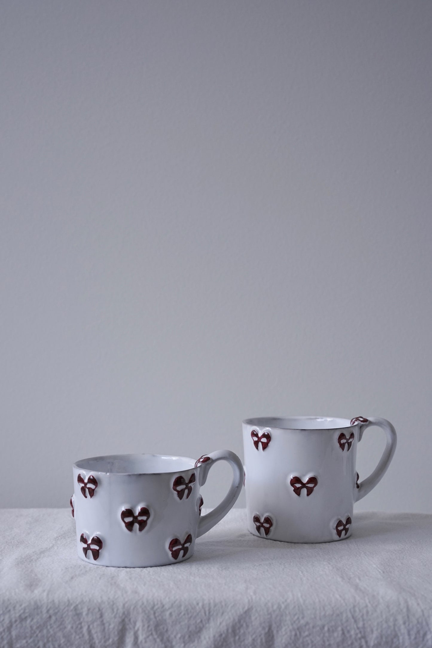 CARRON Paris handmade white ceramic Nœud-Nœud Trianon mug featuring a hand-painted deep bordeaux bow handle detail, in small and medium sizes. 