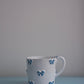 CARRON Paris handmade white ceramic Nœud-Nœud Trianon mug in medium, featuring a hand-painted sky blue bow detail on a blue benchtop.
