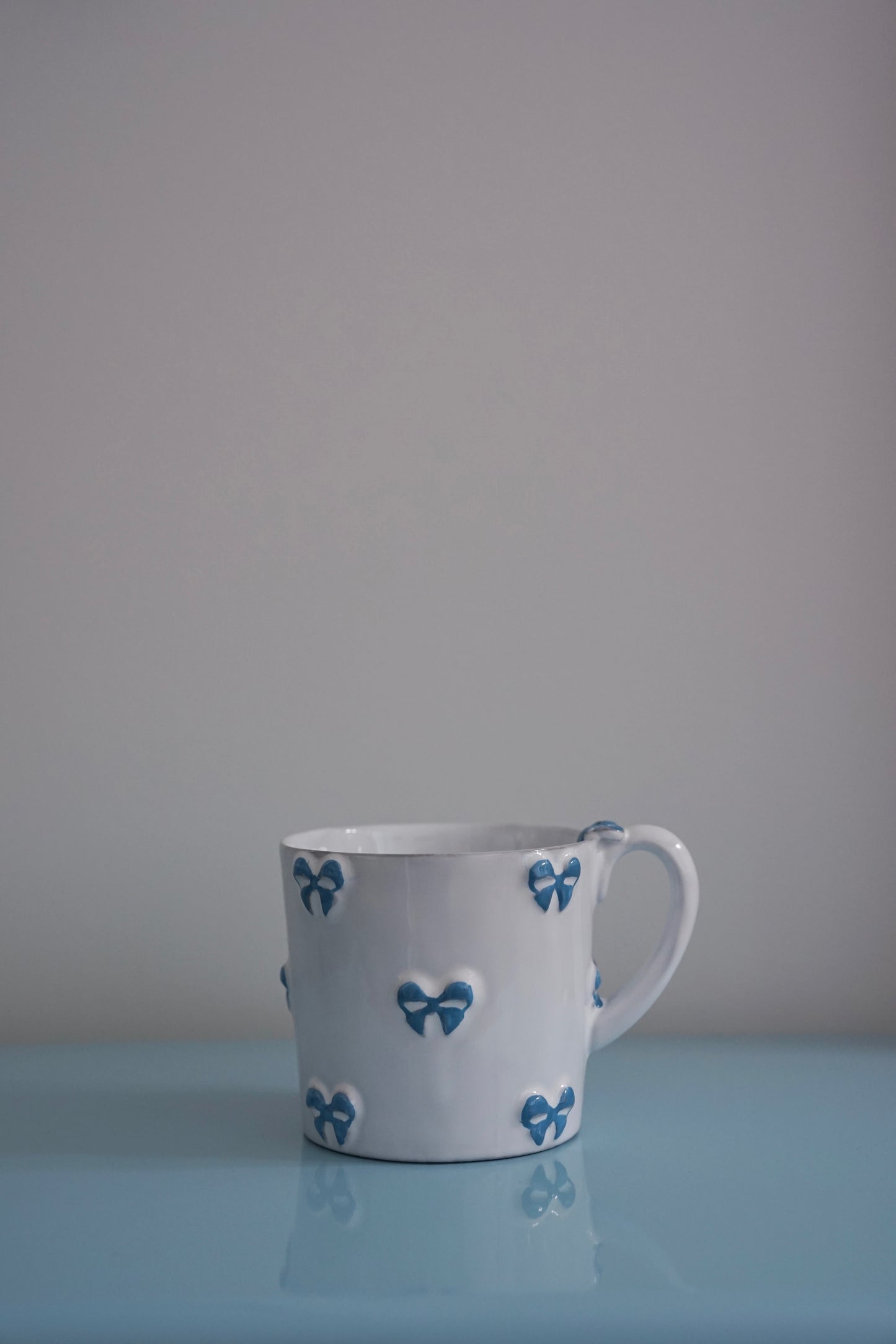CARRON Paris handmade white ceramic Nœud-Nœud Trianon mug in medium, featuring a hand-painted sky blue bow detail on a blue benchtop.