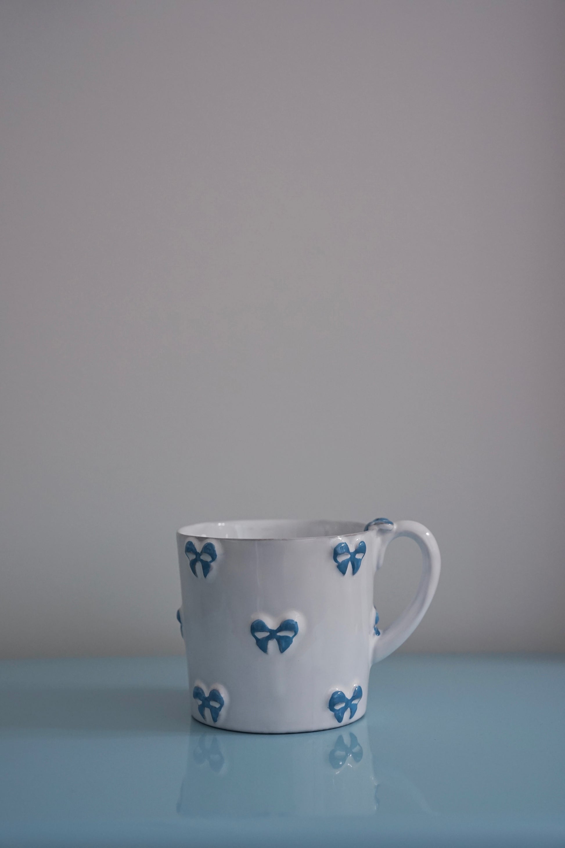 CARRON Paris handmade white ceramic Nœud-Nœud Trianon mug in medium, featuring a hand-painted sky blue bow detail on a blue benchtop.