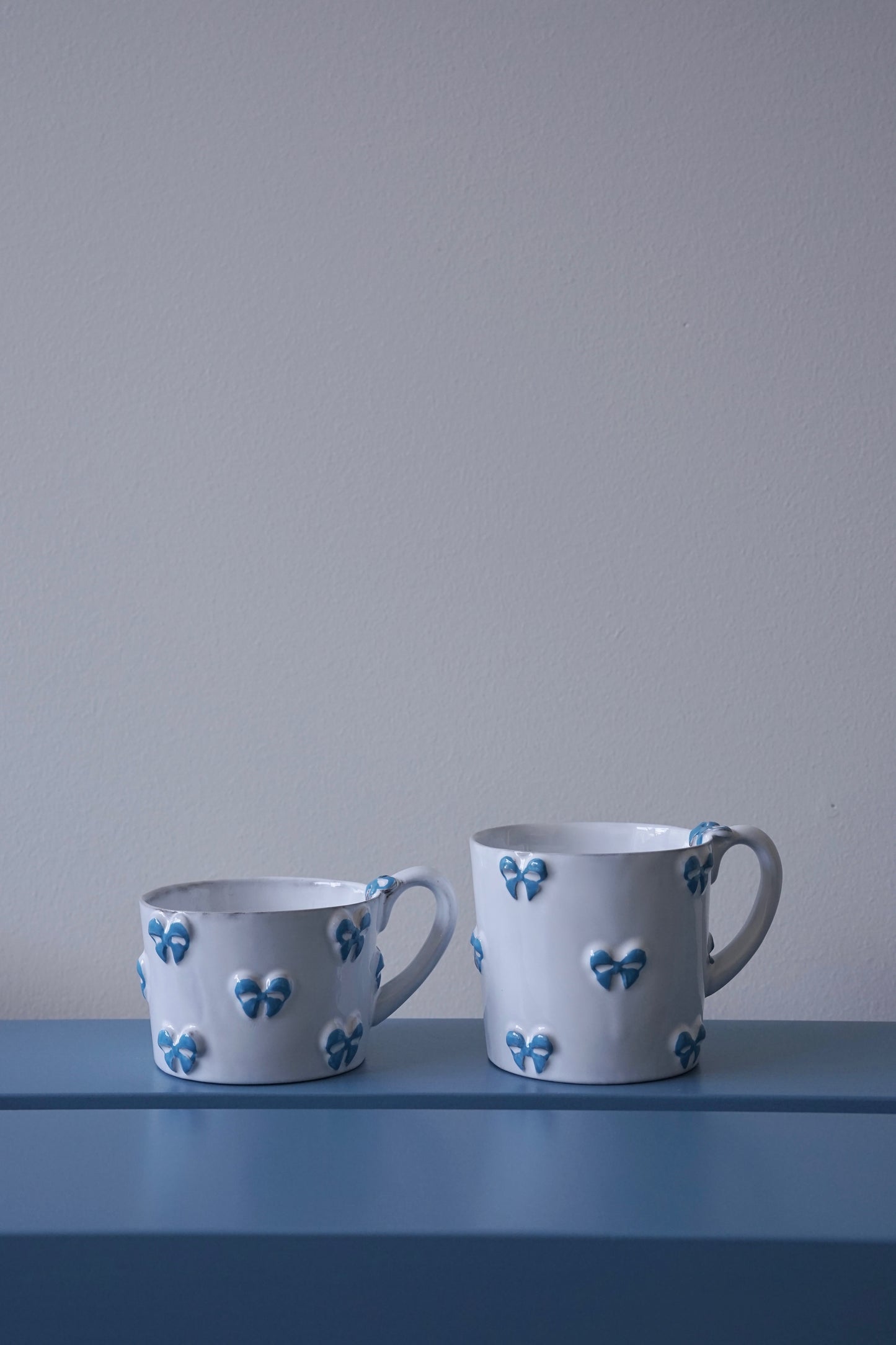 Small and medium CARRON Paris handmade white ceramic Nœud-Nœud Trianon mug in sky blue on a blue bench.