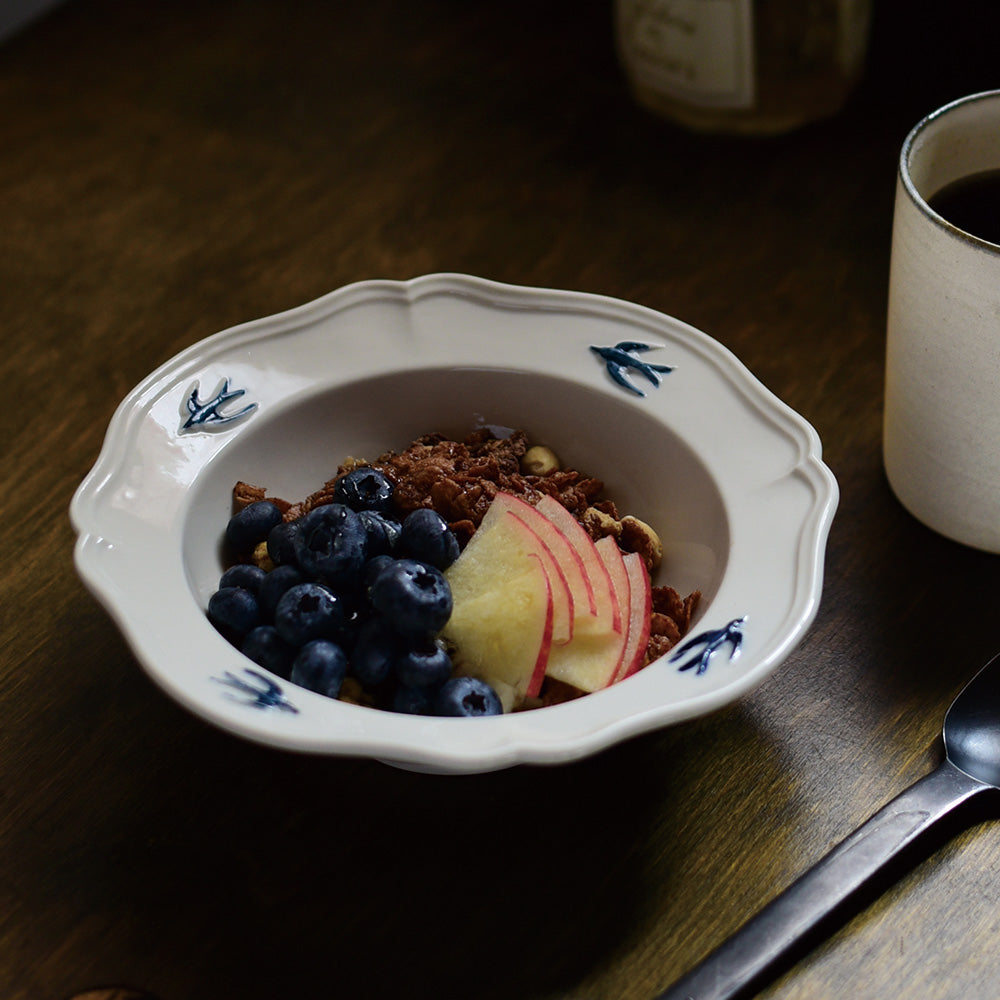 A close-up shot of Studio M' Early Bird 170 porcelain bowl featuring a hand-painted blue swallow relief on a scalloped floral rim, used as a cereal bowl. 