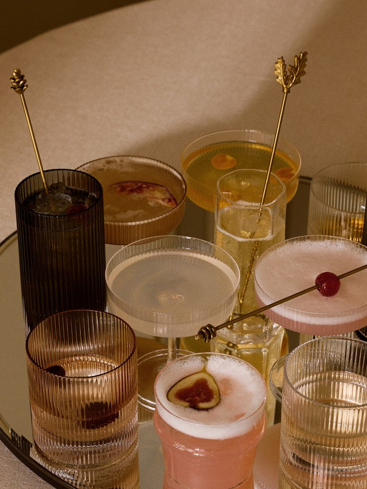 An assortment of cocktails in various ferm LIVING Ripple glassware, including champagne glasses, tumblers, and highball glasses, arranged on a mirrored tray against a warm beige background.