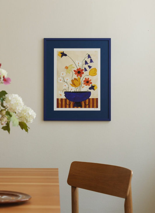 a lifestyle photo of the art print Colourful Bunch in Blue Vase by Frankie Penwill, exclusively created for The Poster Club, in a blue wooden frame with a blue paper cutout. 