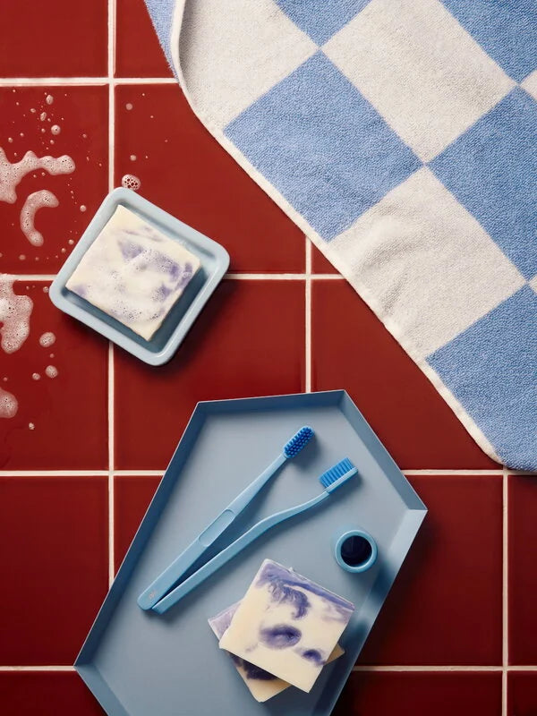 HAY Tann toothbrushes blue on a blue metal tray laid on burgundy tiles next to blue soap dish.