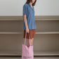 A woman stands in a minimalist room with beige shelves, wearing a blue short-sleeve button-up shirt, holding HÔTEL827 Sun Kissed pink canvas tote bag.