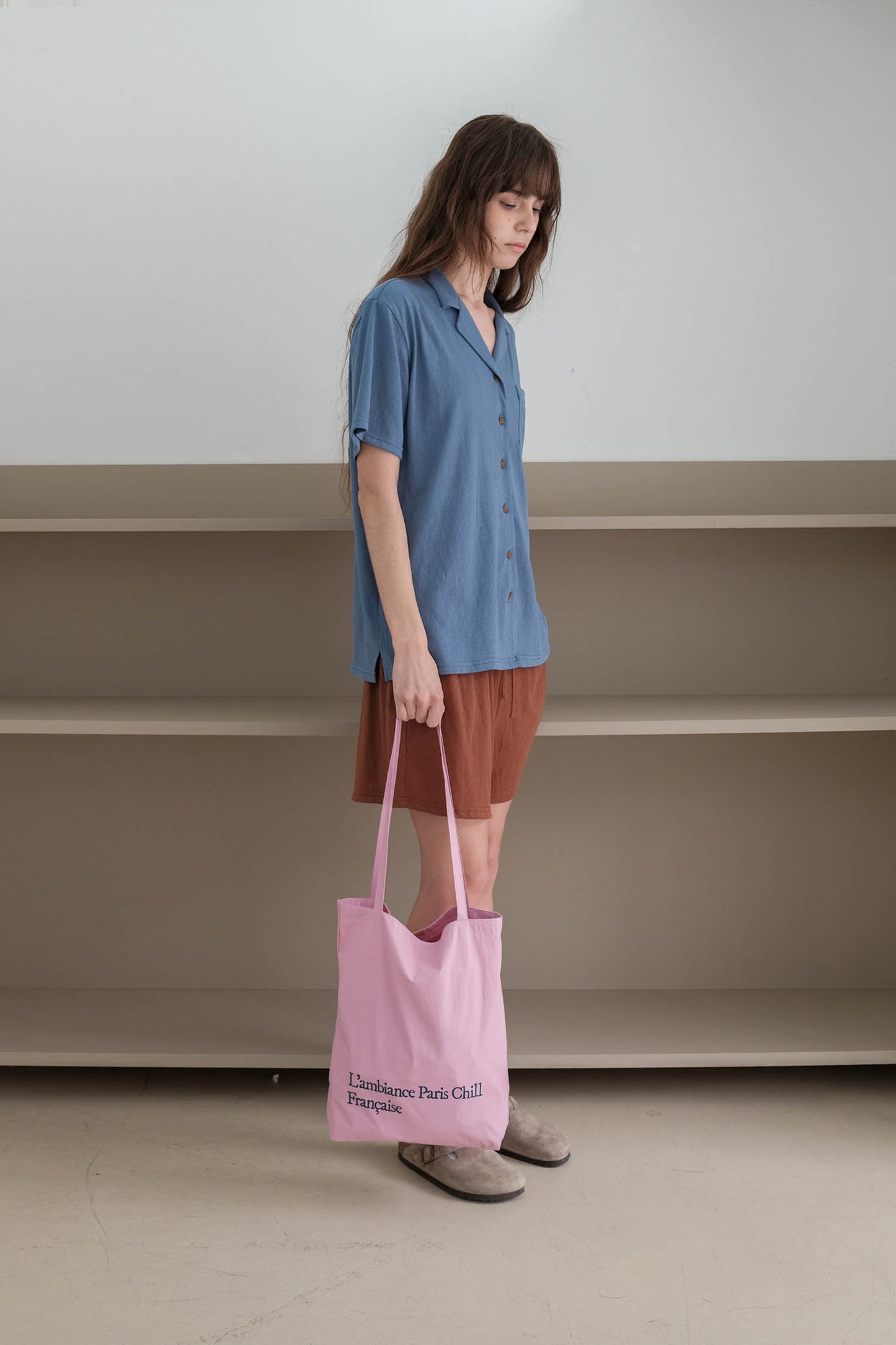 A woman stands in a minimalist room with beige shelves, wearing a blue short-sleeve button-up shirt, holding HÔTEL827 Sun Kissed pink canvas tote bag.