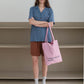 A woman stands with arms crossed, wearing a blue short-sleeve button-up shirt, holding HÔTEL827 Sun Kissed pink canvas tote bag.