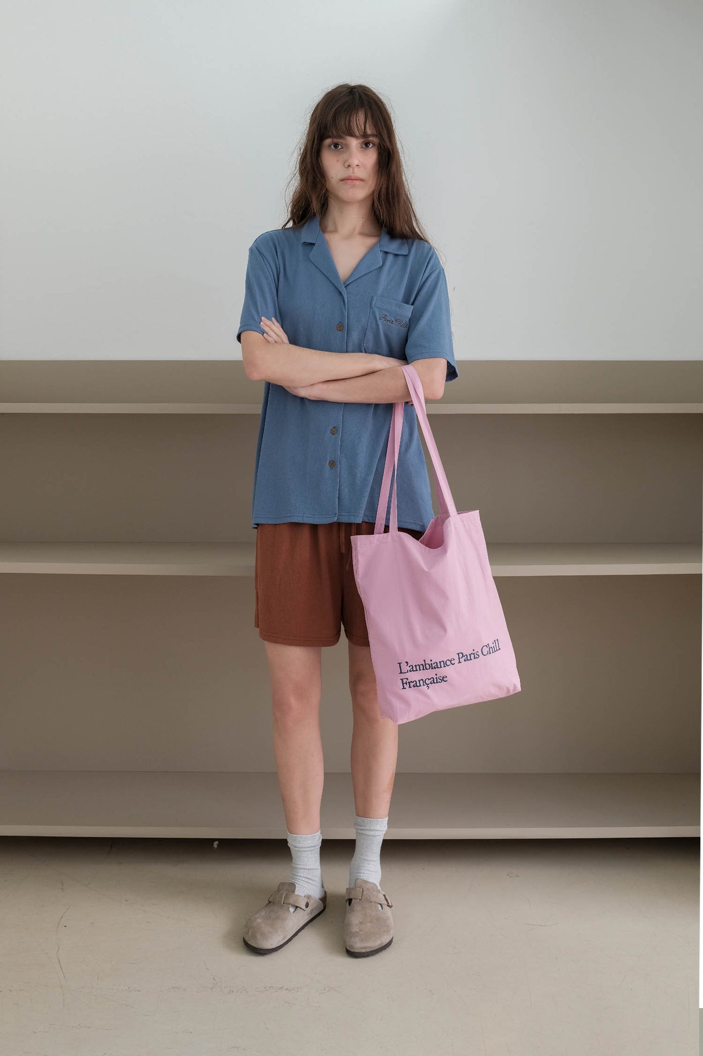 A woman stands with arms crossed, wearing a blue short-sleeve button-up shirt, holding HÔTEL827 Sun Kissed pink canvas tote bag.