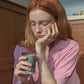 A woman with red hair sits on a wooden floor in a kitchen, wearing a pink short-sleeve top and holding Hotel827 Sun Kissed Glass Cup in Smokey Grey.