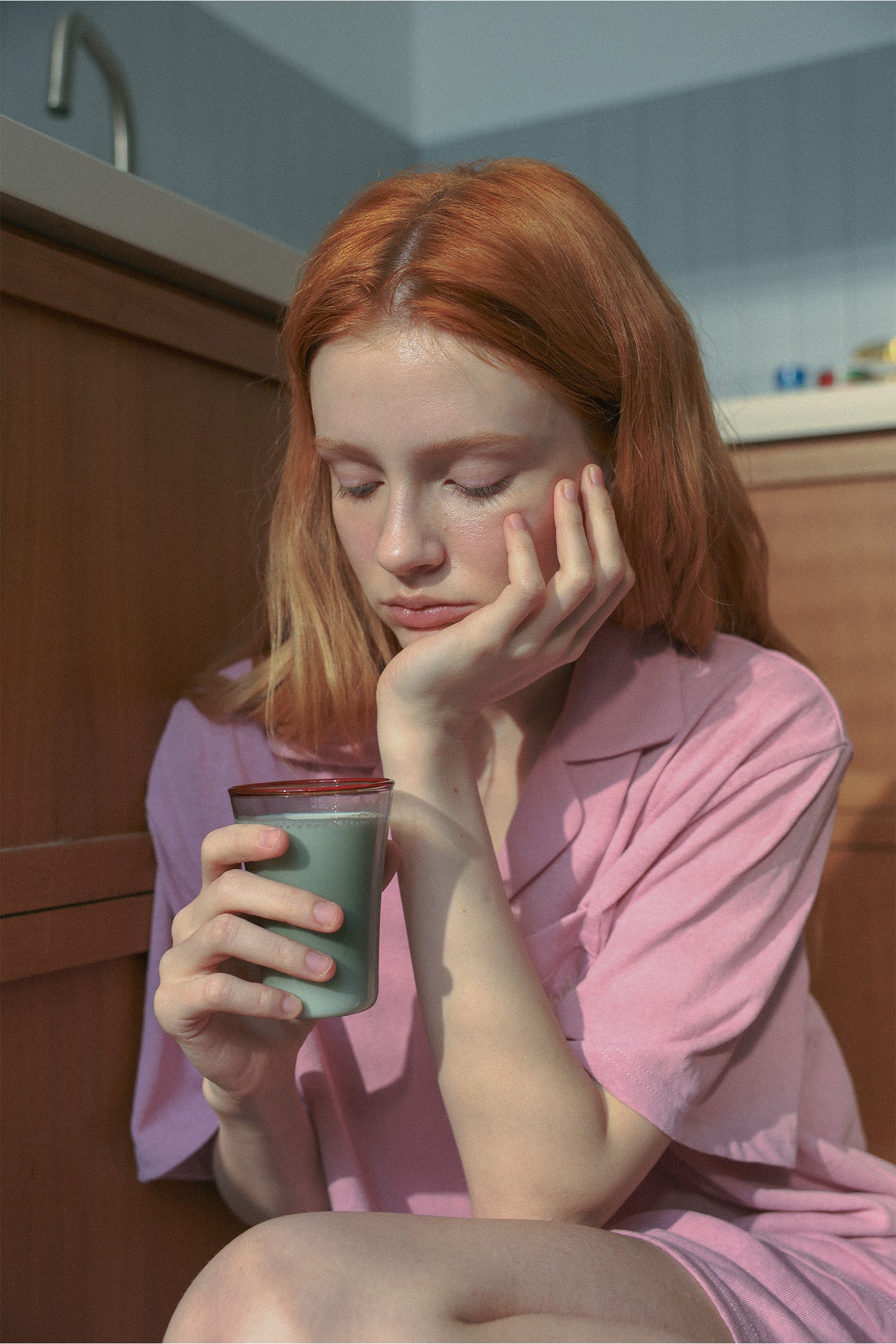 A woman with red hair sits on a wooden floor in a kitchen, wearing a pink short-sleeve top and holding Hotel827 Sun Kissed Glass Cup in Smokey Grey.