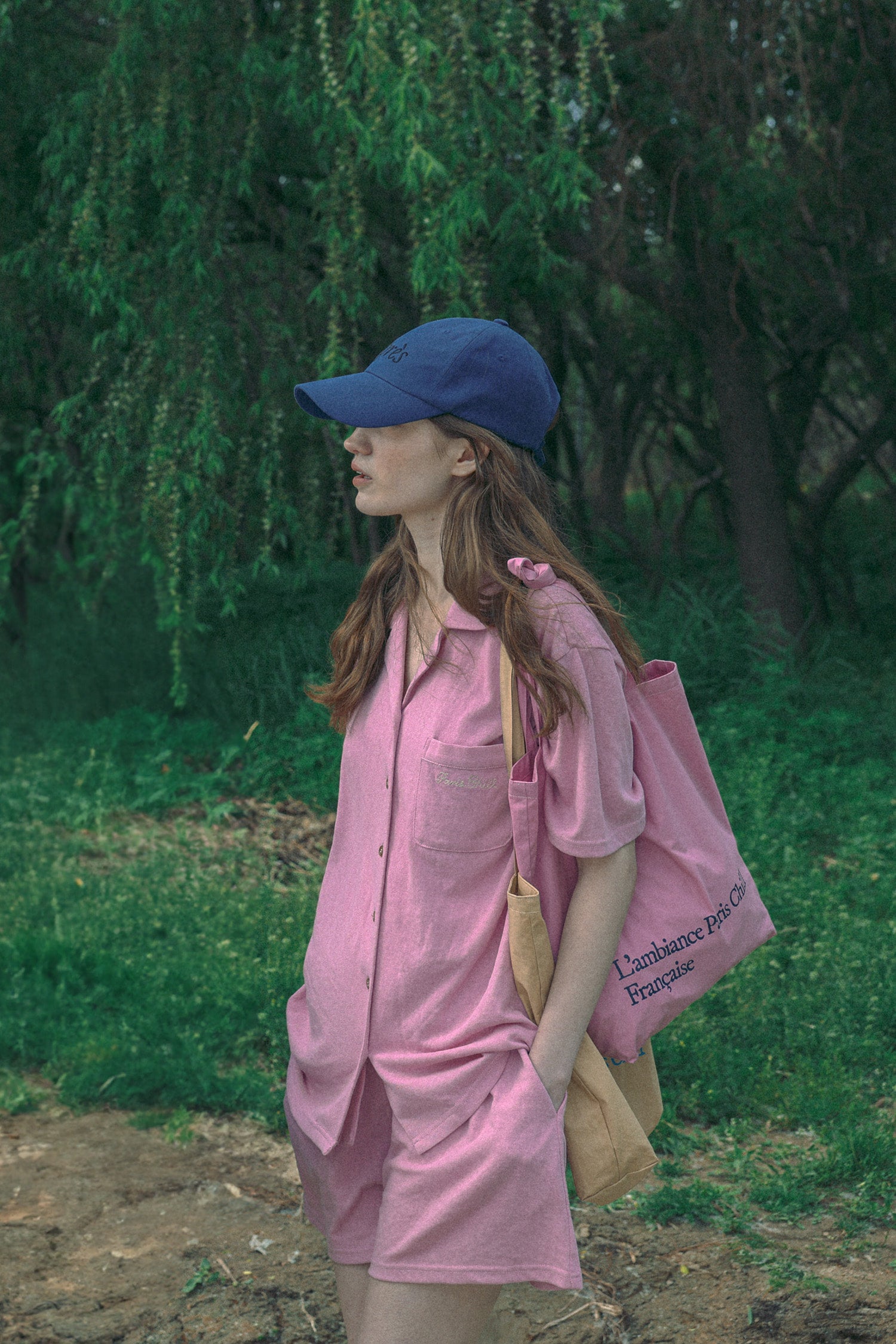 A woman wearing a pink casual wear set, wearing HÔTEL827 Sun Kissed pink canvas tote bag over shoulder with Hotel827 sun kissed blue baseball cap.