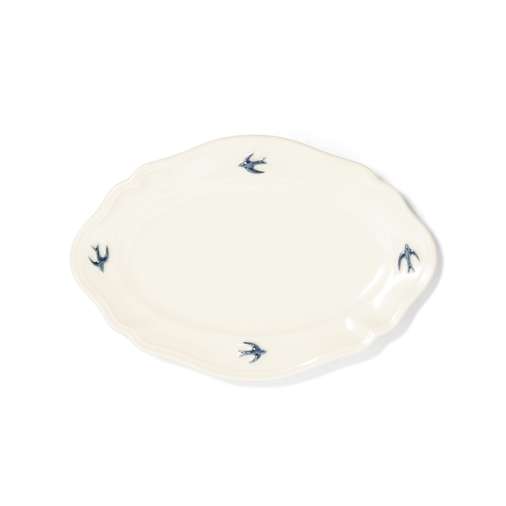 Studio M' Early Bird Oval Plate L in white porcelain, featuring a scalloped rim and a hand-painted blue swallow relief. A shot taken from the aerial angle.