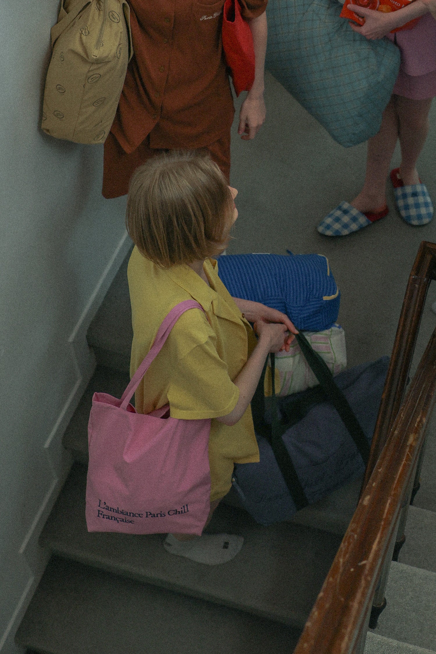 A woman wearing a yellow pyjamas, wearing HÔTEL827 Sun Kissed pink canvas tote bag over shoulder and holding Hotel827 travel pouches.